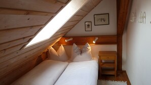 Een knus zolderkamer met een schuine kap, houten balken, een bed met witte lakens, een nachtkastje en twee wandlampen.