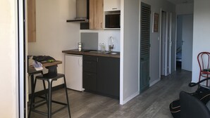 Pieni ja käytännöllinen keittiö, jossa on pesuallas, työtaso ja pieni jääkaappi. Etualalla on baarituoli ja tuoli.