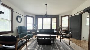 Un salon comprenant un canapé, des fauteuils, une table basse et un tapis. On y trouve des fenêtres équipées de stores, une horloge au mur et un lustre.