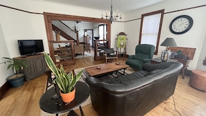 Un salon avec un canapé en cuir noir, une table basse en bois, un téléviseur posé sur un meuble, un escalier et une horloge au mur.