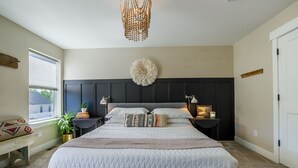 Une chambre à coucher avec un grand lit, une tête de lit sombre, des tables de chevet avec des lampes, un luminaire suspendu et une fenêtre avec des stores.