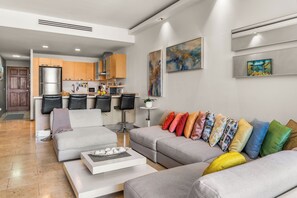 A modern living room with a grey sectional sofa, a coffee table, and a kitchen area with wooden cabinets and a stainless steel refrigerator.