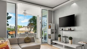 Een moderne woonkamer met een grote televisie, witte meubels en een balkon met uitzicht op palmbomen en gebouwen.