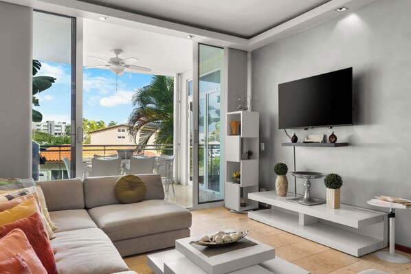 A modern living room with a large TV, white furniture, and a balcony with a view of palm trees and buildings.