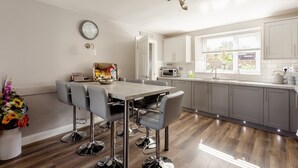 A modern kitchen with grey cabinets, a dining table with chairs, a clock, and a window with a view of greenery. - Neds Brae House (Ballymena)