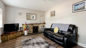 A living room with a fireplace, a black leather sofa, a flat-screen TV, and a stone fireplace surround. - Neds Brae House (Ballymena)