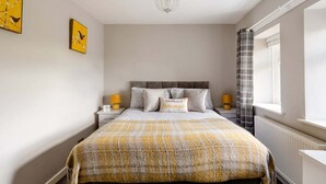 A bedroom with a bed, two bedside tables with lamps, a window with curtains, and artwork on the wall. - Neds Brae House (Ballymena)