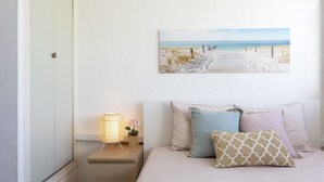 A bedroom with a bed, pillows, a nightstand with a lamp, and a beach scene painting on the wall. - Holidays in MARINA in PORT CAMARGUE and 100m from the beach! (Le Grau-du-Roi)