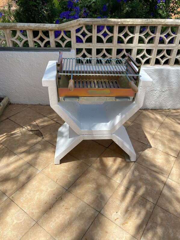 A barbecue grill with a metal grate and a handle, set on a tiled patio with a white wall and decorative fence in the background. - Traditional family villa, private pool and hillside views, 5 mins to Moraira. (Teulada)