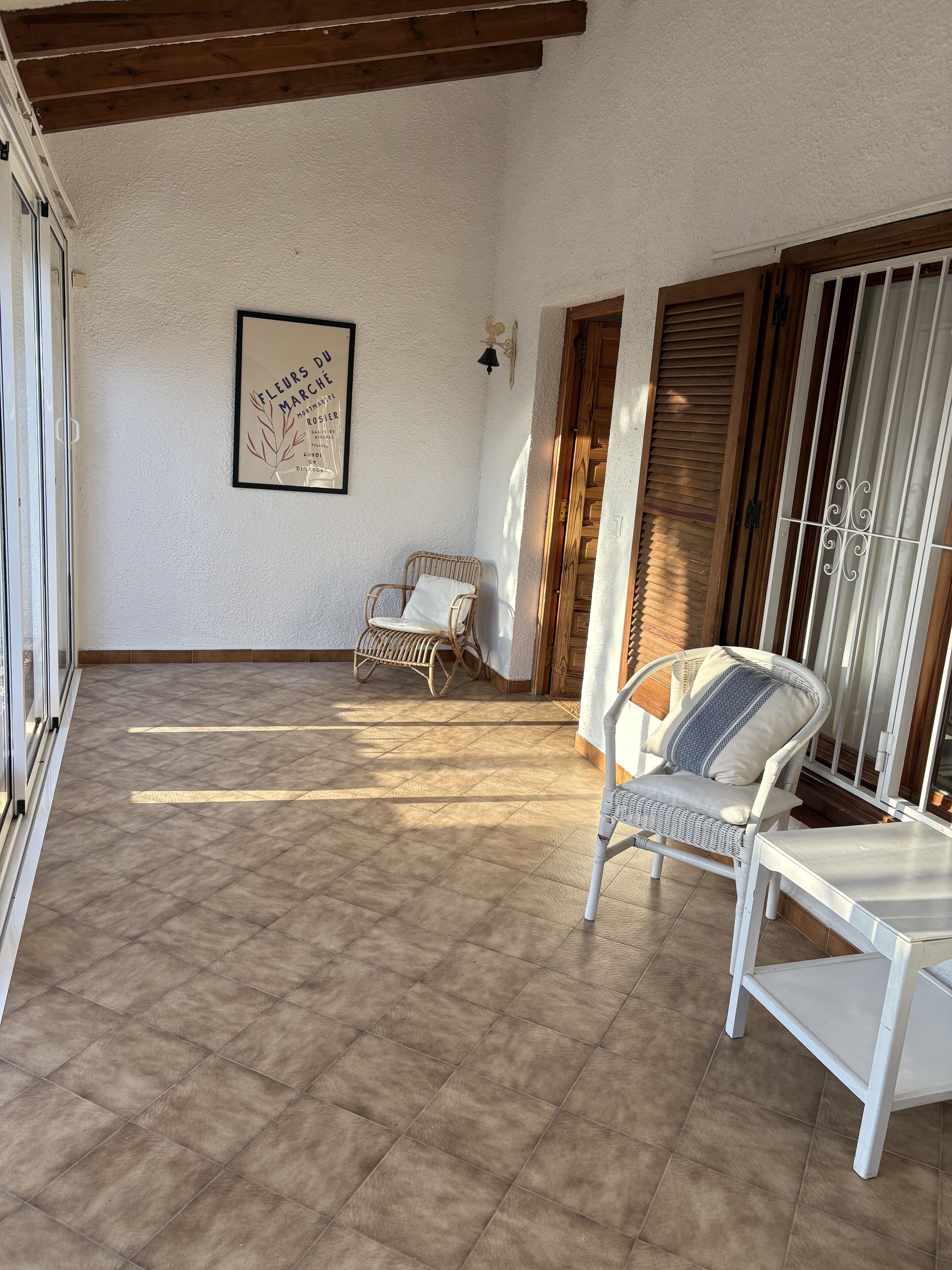 A covered patio area with a tiled floor, two wicker chairs, a white table, and a framed sign on the wall.