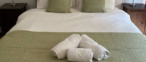 A neatly made bed with a green patterned bedspread, two pillows with a geometric design, and a pair of folded towels on the bed.