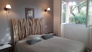 Un dormitorio con una cama, dos mesitas de noche, una lámpara de pared, un cuadro enmarcado y una ventana con vistas a la vegetación.