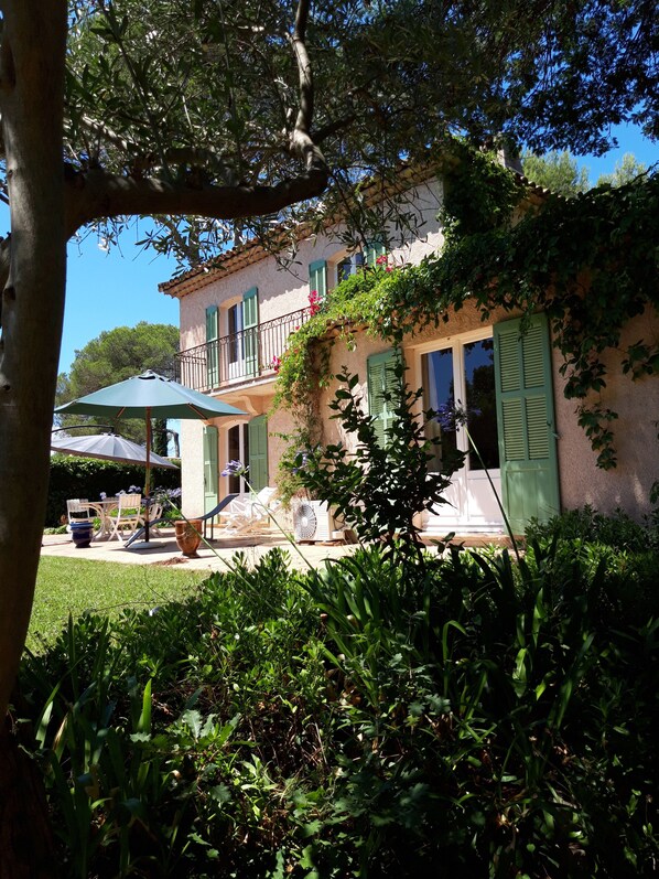 A two-story building with green shutters, a balcony, and a garden area with outdoor furniture.