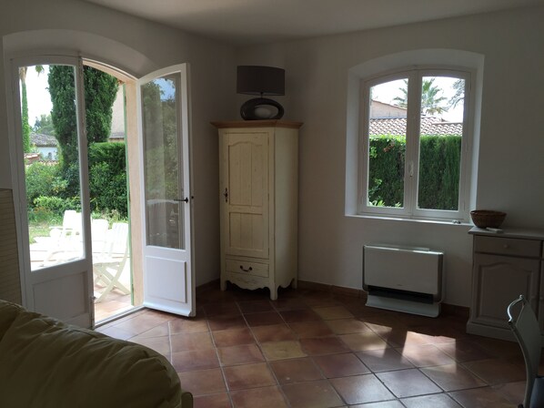 A room with a sliding glass door leading to a garden, a white cabinet with a radio on top, a window with a view of greenery, and a radiator on the floor.