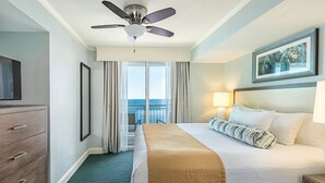 A hotel room with a bed, a ceiling fan, a TV, a nightstand, a lamp, and a view of the ocean. - One-Bedroom Oceanfront Condo on Myrtle Beach (North Myrtle Beach)