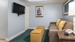 A hotel room with a flat-screen TV mounted on the wall, a framed picture, a brown sofa with cushions, a small ottoman, and a bedside table with a phone and a lamp. - One-Bedroom Oceanfront Condo on Myrtle Beach (North Myrtle Beach)