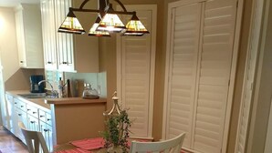 Un coin repas avec une table en bois, des chaises blanches et un banc agrémenté d’un coussin rouge. Un lustre est suspendu au-dessus de la table, et une petite plante y est posée.