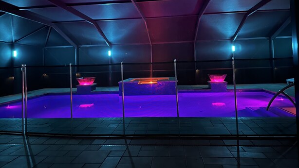 Zona de piscina interior moderna, con poca luz, diseño geométrico en el techo e iluminación púrpura.