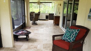 Un patio techado con una mesa y sillas de comedor, una silla de mimbre con cojín rojo, y piso de baldosas.