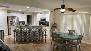 Una cocina moderna con una isla central, taburetes de bar, una mesa de comedor con sillas y un ventilador de techo.