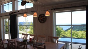 A dining area with a wooden table and chairs, large windows with a view of the outdoors, pendant lights, and a wall clock.