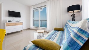 Un salon moderne avec un canapé, un téléviseur, une petite table et une vue sur la mer, visible à travers une grande fenêtre.