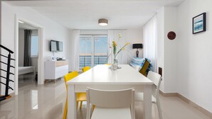 Un espace repas moderne avec une table blanche et des chaises jaunes, un petit meuble blanc et une vue sur la mer grâce à de grandes fenêtres.