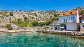 Un bâtiment situé sur la côte, doté d’un balcon donnant sur des eaux turquoise et limpides, d’un mur en pierre et surplombant un paysage vallonné.