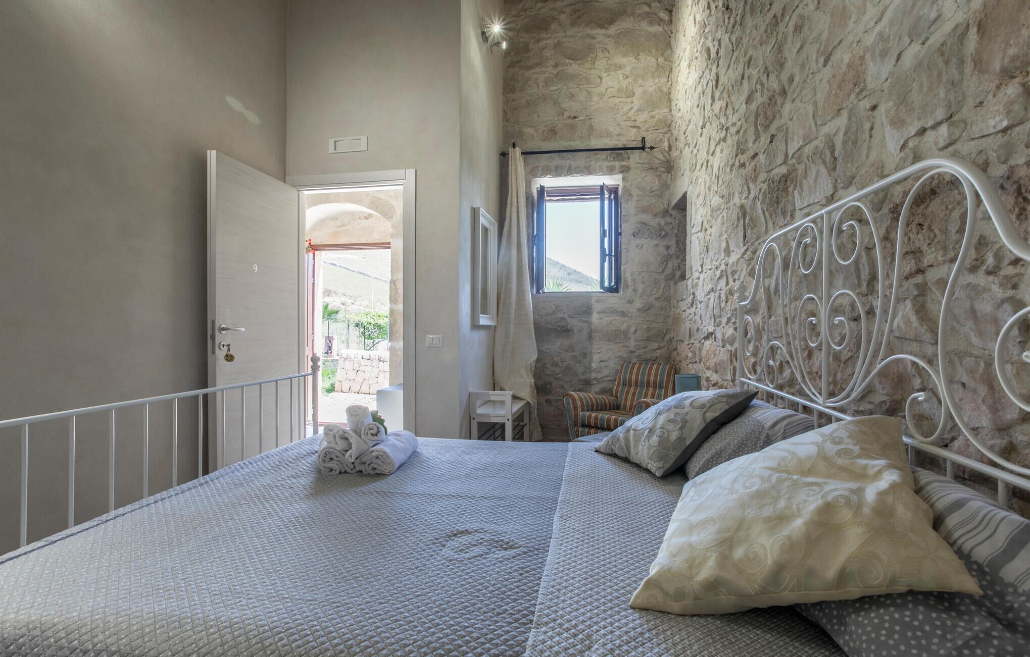 Ein Schlafzimmer mit Steinwand, Metallbettgestell und Blick ins Freie durch ein Fenster.