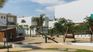 Ein Spielplatz mit Schaukeln und Rutsche, umgeben von Palmen und modernen Gebäuden.