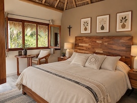 Un dormitorio con cama, mesitas de noche, una silla, una ventana con vista a la vegetación y cuadros en la pared.