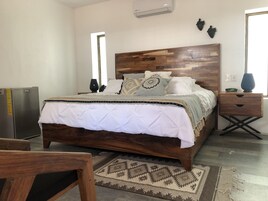 Un dormitorio con cabecera de madera, cama con ropa de cama blanca, mesita de noche con un reloj y aire acondicionado instalado en la pared.