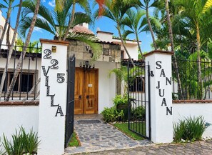 A gated entrance with a stone pathway leading to a wooden door, surrounded by lush greenery and palm trees.