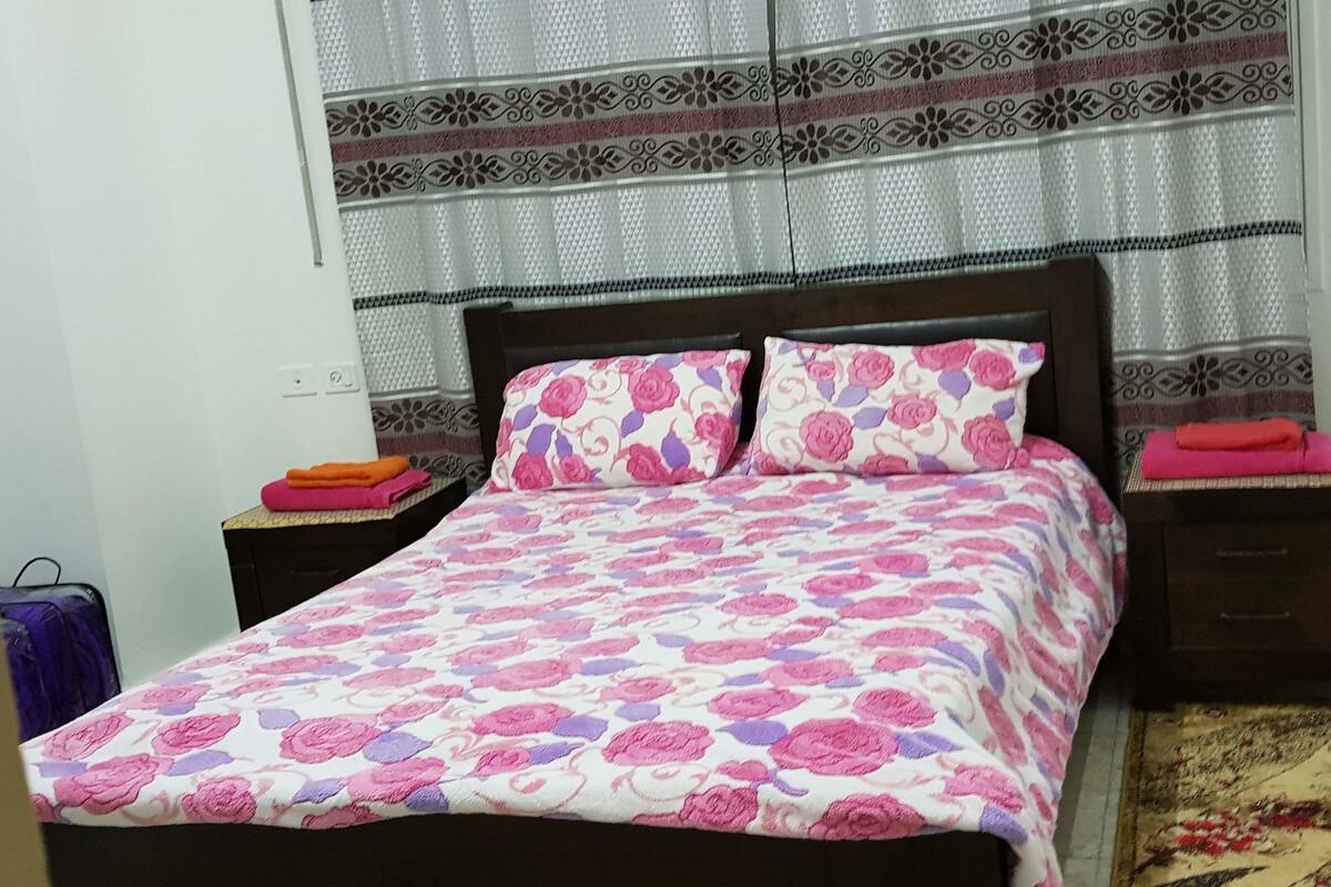 A bed with a floral bedspread, two pillows, a nightstand with a lamp, and a suitcase.
