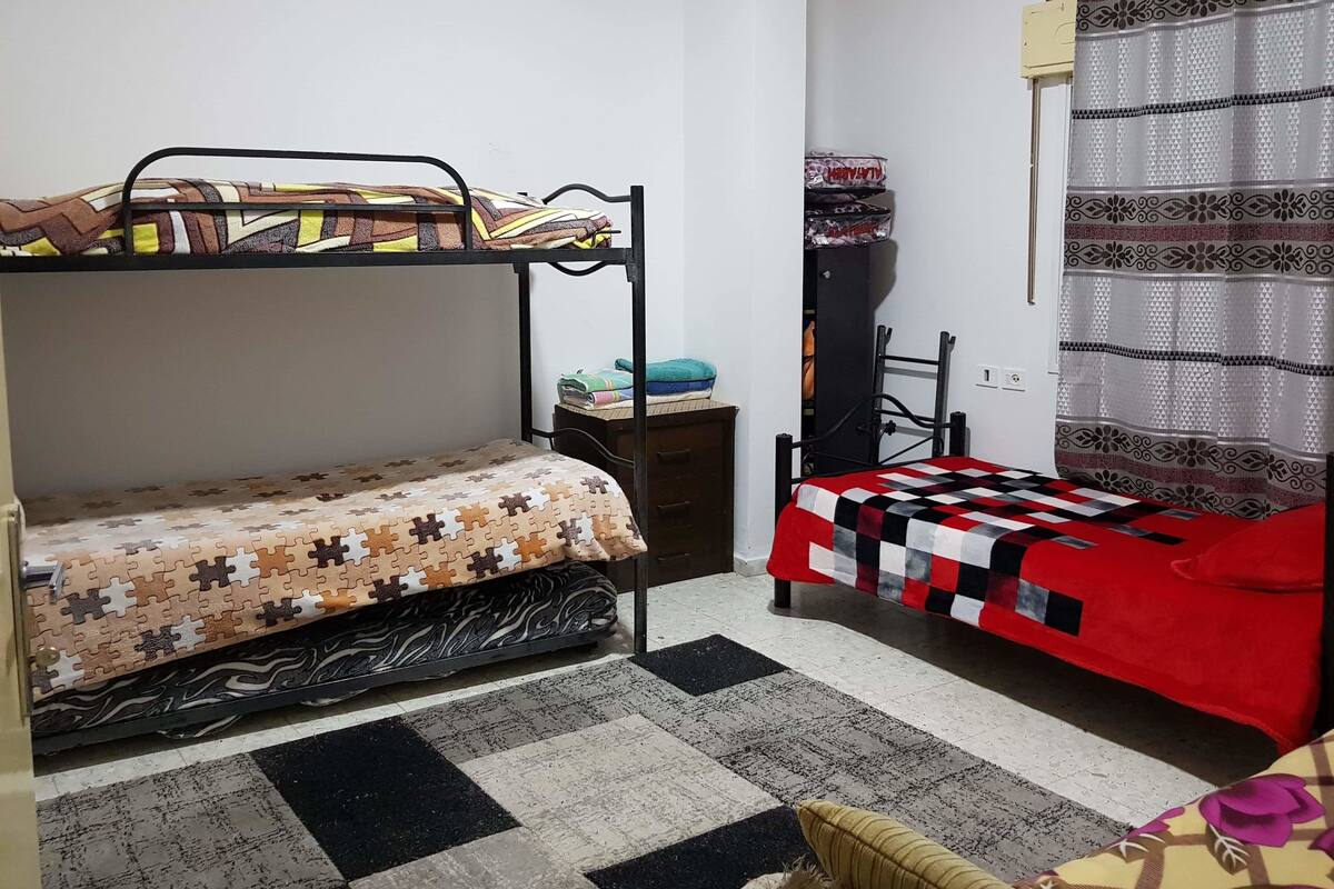 A dormitory room with two bunk beds, a small table, a chair, and a patterned rug.