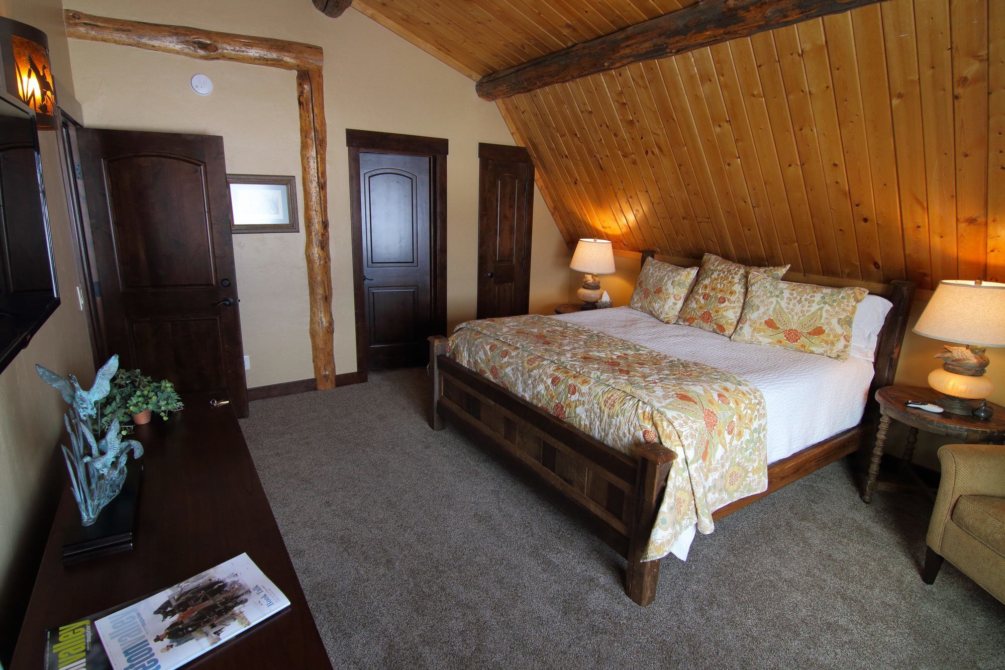 Ein Schlafzimmer mit Holzboden, einem Bett mit Blumenmuster-Bettwäsche, einem Nachttisch mit Lampe und einem Schreibtisch mit einer Zeitschrift.