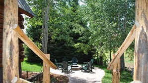 A wooden deck with a fire pit and Adirondack chairs, surrounded by lush greenery.