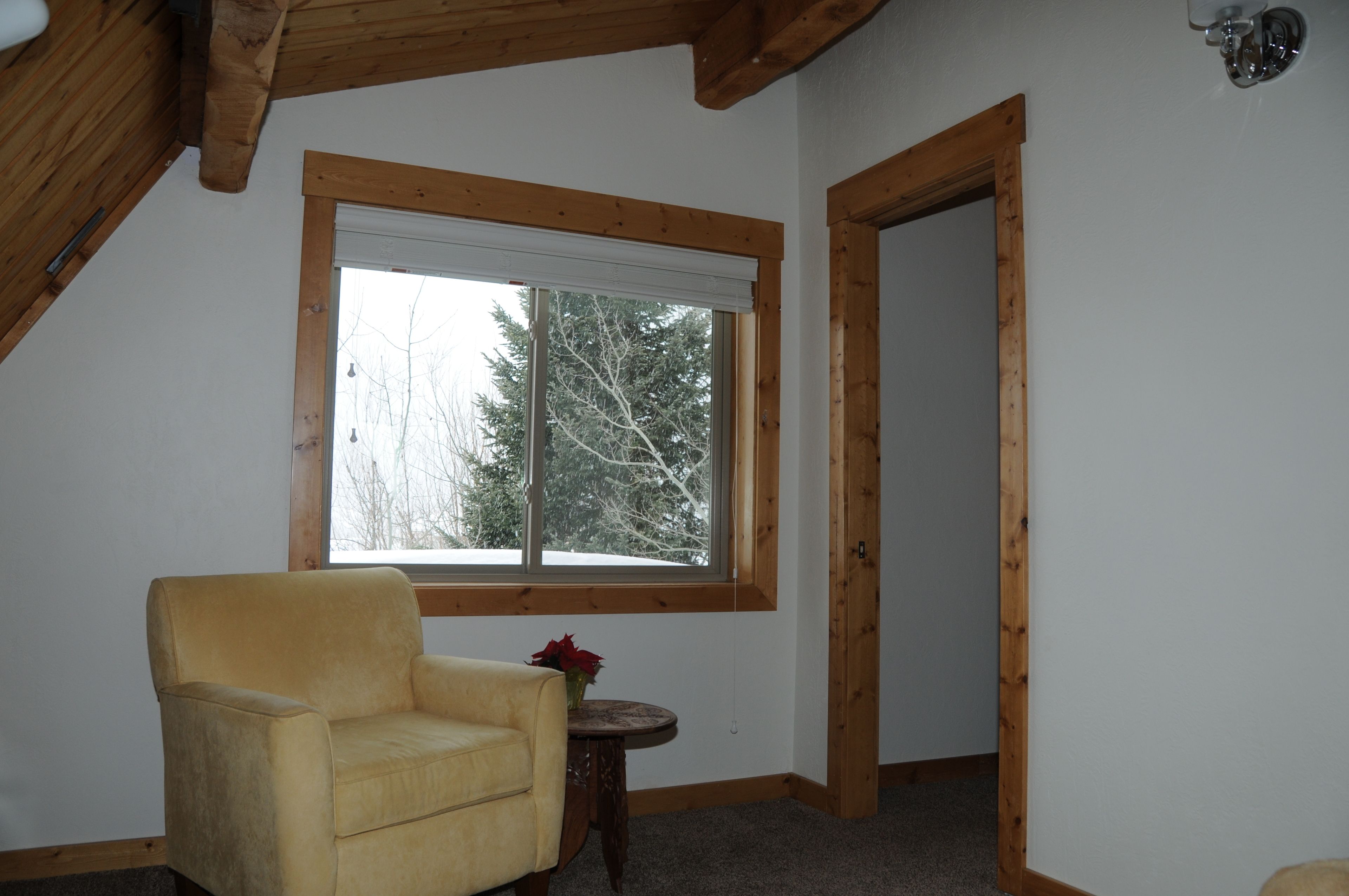 A room with a large window, a yellow armchair, a small round table with a vase, and wooden beams on the ceiling.