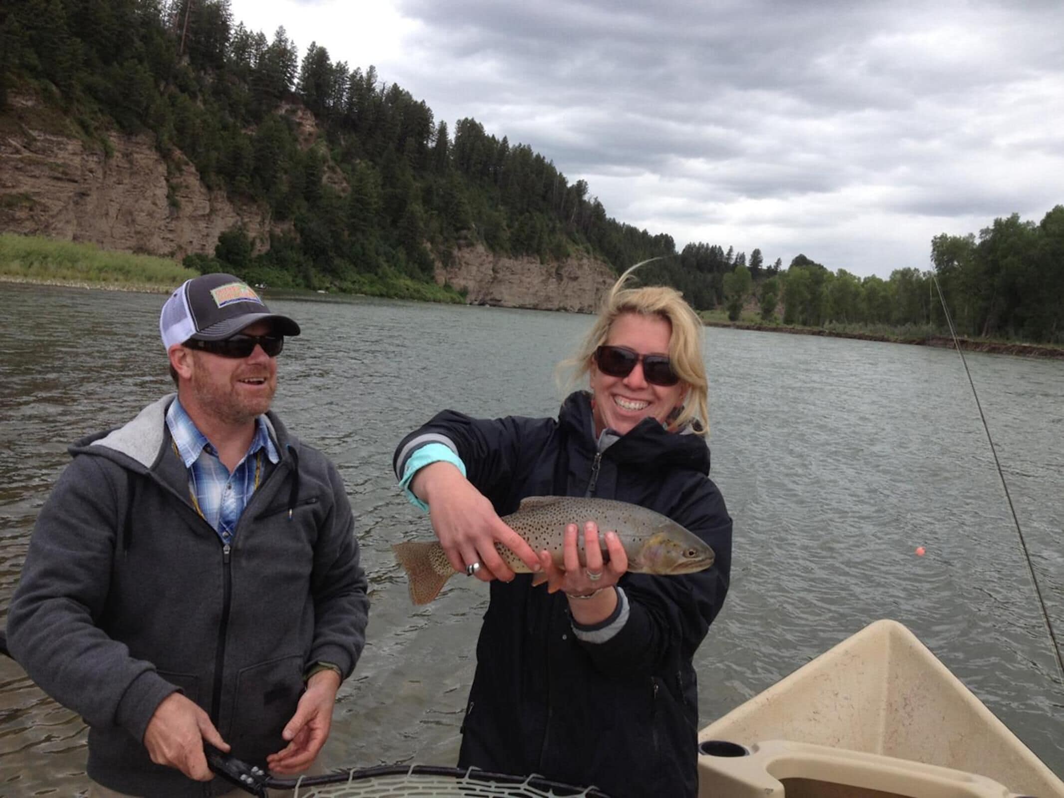 Two people holding a fish by a river with a boat in the foreground.