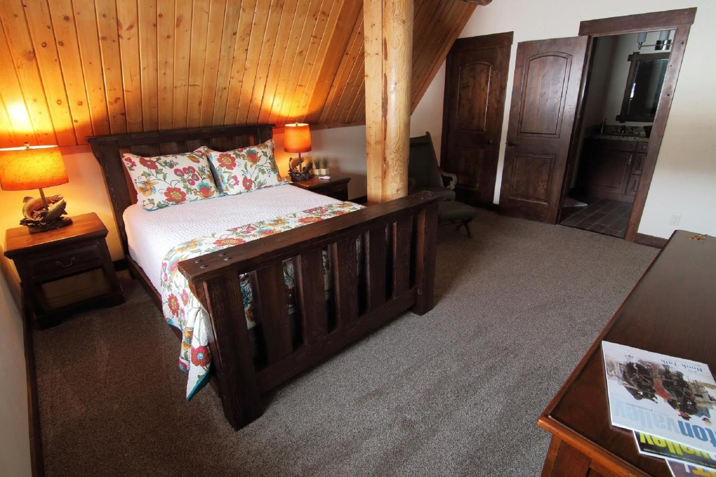 Un dormitorio con techo de madera, una cama con ropa de cama floral, dos lámparas de noche, una mesita de noche de madera y un aparador de madera.