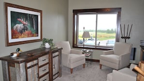 A room with a wooden desk, two armchairs, a lamp, and a window with a view of the outdoors.