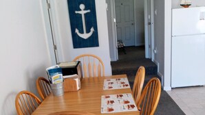 A dining area with a wooden table and chairs, a blue anchor wall decoration, and a kitchen area with a white refrigerator. - Orchard Lane at Round Lake. (Manitou Beach)