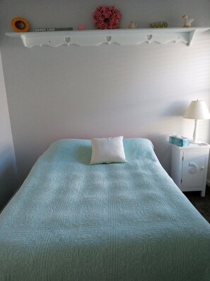 A bed with a textured blue cover, a white pillow, a small nightstand with a lamp, and a shelf with decorative items.