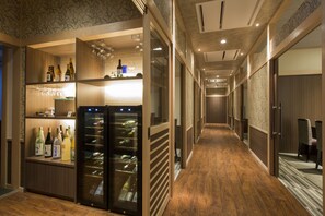 A hotel corridor with wooden flooring, a wine refrigerator, and shelves with bottles and glasses.