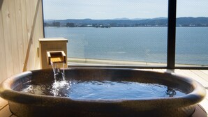 A stone hot tub with water flowing from a faucet, positioned next to a wooden wall and a large window overlooking a body of water and distant hills.