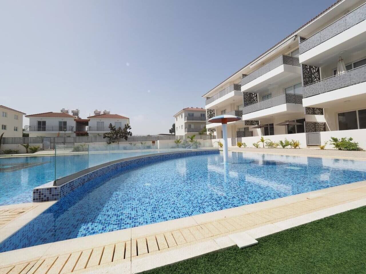 A swimming pool with a tiled edge, surrounded by a grassy area and modern buildings with balconies.