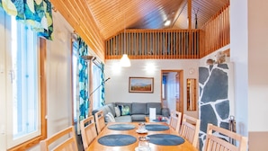 A spacious dining area with a wooden table and chairs, a high ceiling with wooden beams, and a view of a cozy living space in the background.