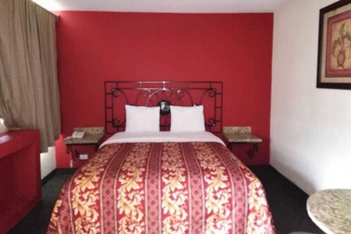 Une chambre d’hôtel avec un mur rouge, un cadre de lit en métal, une couvre-lit à motifs, une table de chevet et une petite table ronde.