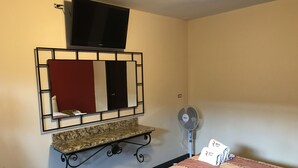 Habitación de hotel con televisor montado en la pared, tocador con espejo, ventilador y cama con colcha a rayas.