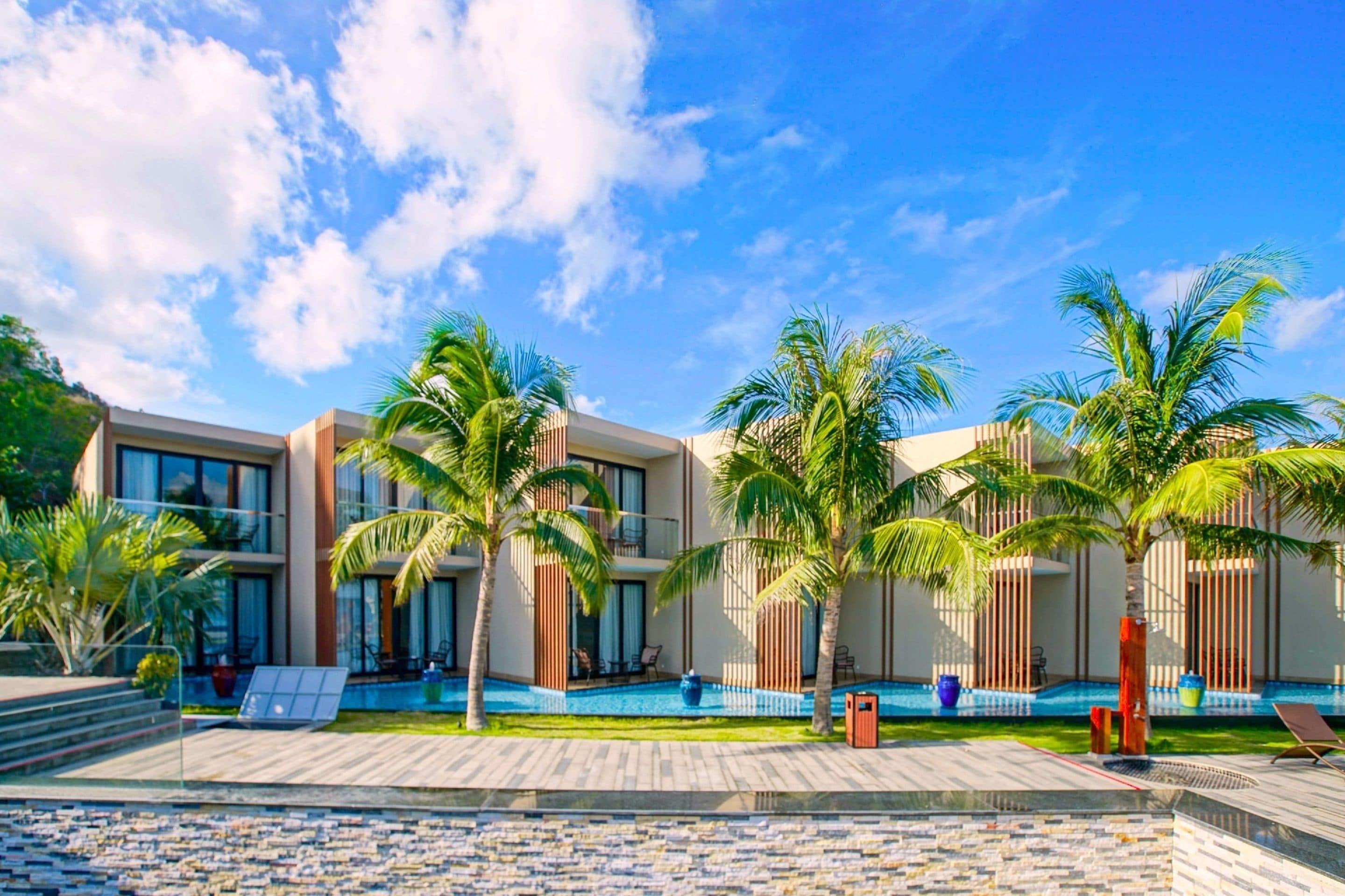 A modern hotel with a pool area, palm trees, and a clear blue sky.